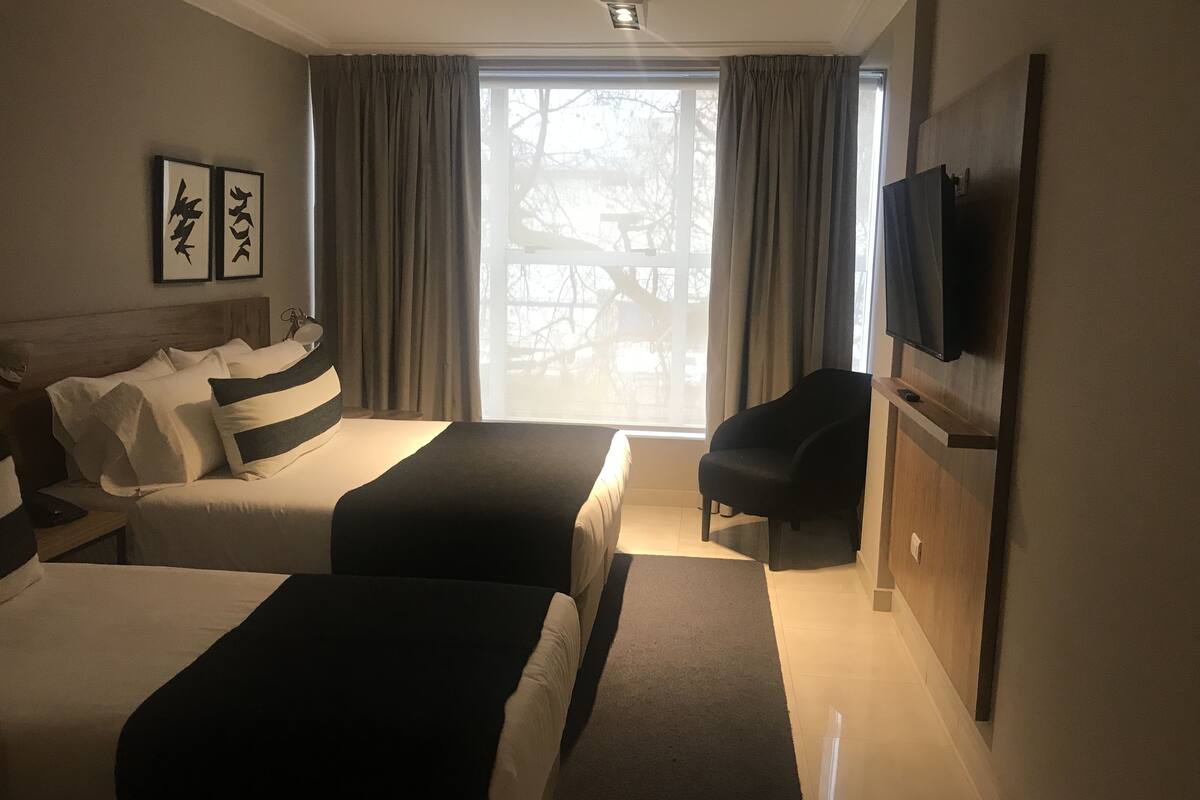 Ett hotellrum med två sängar, en platt-TV, en stol och ett fönster med gardiner.