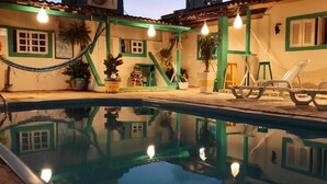 Una piscina con sillones de descanso y plantas en macetas, rodeada por un edificio con detalles verdes y ventanas.