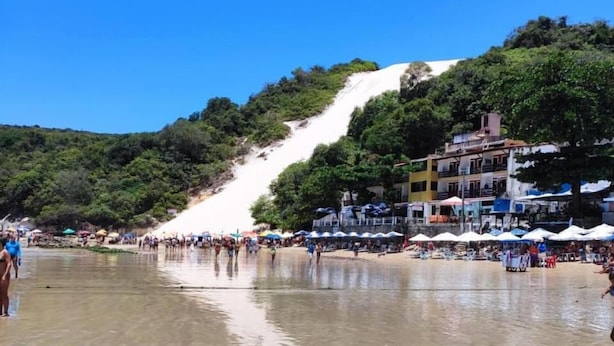 Una playa con una pendiente arenosa que lleva a una colina, rodeada de edificios y sombrillas.