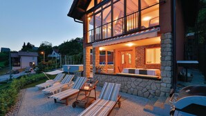 Ein modernes Haus mit Steinverkleidung, großen Fenstern und einer Terrasse mit Liegestühlen und einem Tisch.