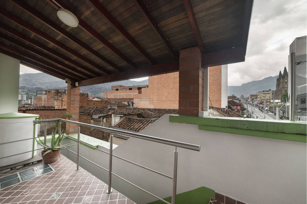 Een balkon met een bloempot, metalen hekwerk en uitzicht op een stadsgezicht met gebouwen en bergen op de achtergrond.