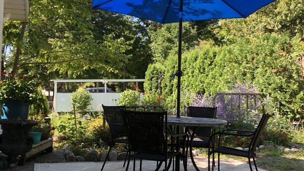 An outdoor patio with a table and chairs under a blue umbrella, surrounded by lush greenery and a stone floor. - Walk everywhere in the adorable village of Suttons Bay. (SUTTONS BAY)
