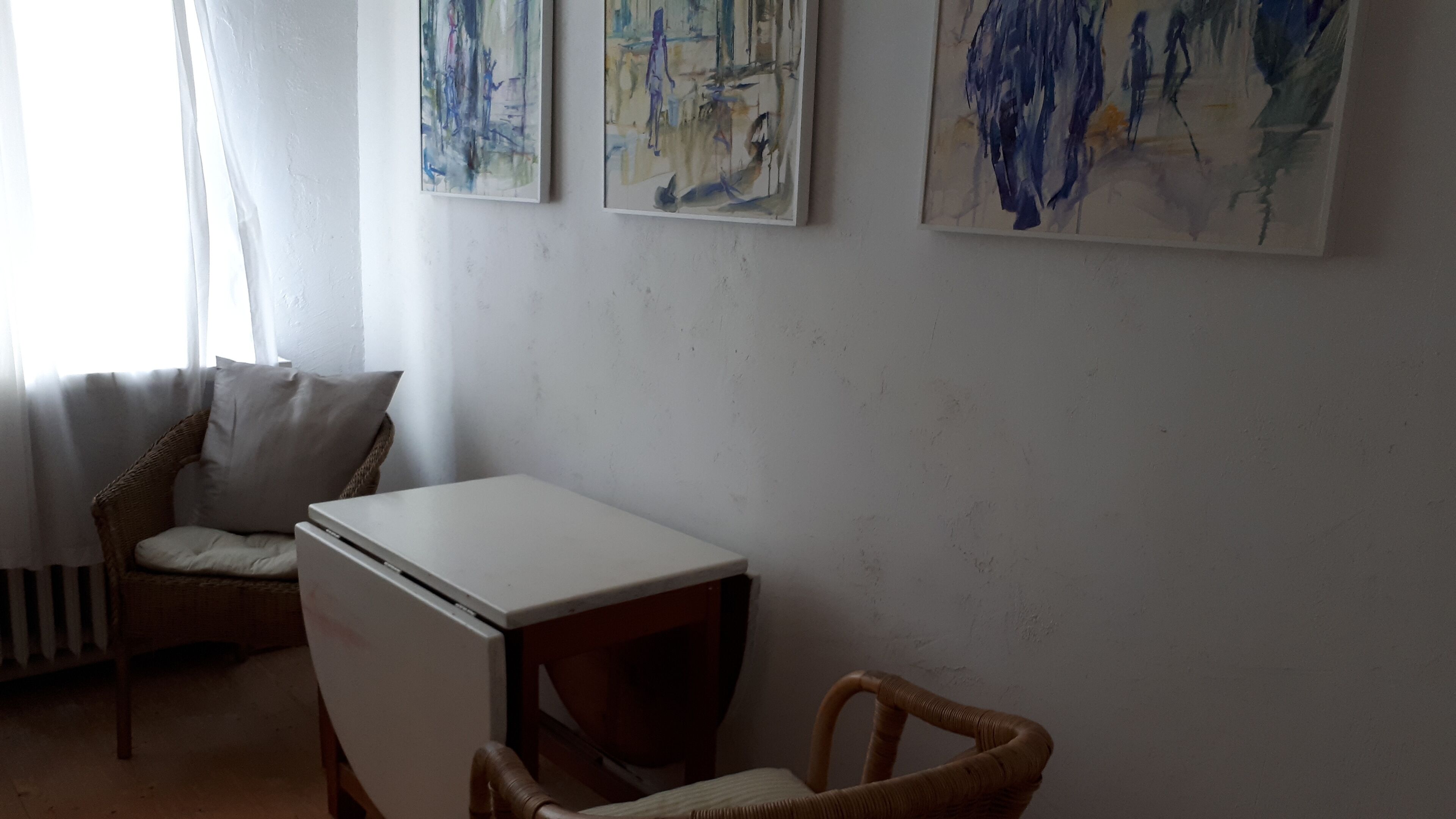 A room with a white table, a wicker chair, a white sofa, and three abstract paintings on the wall.