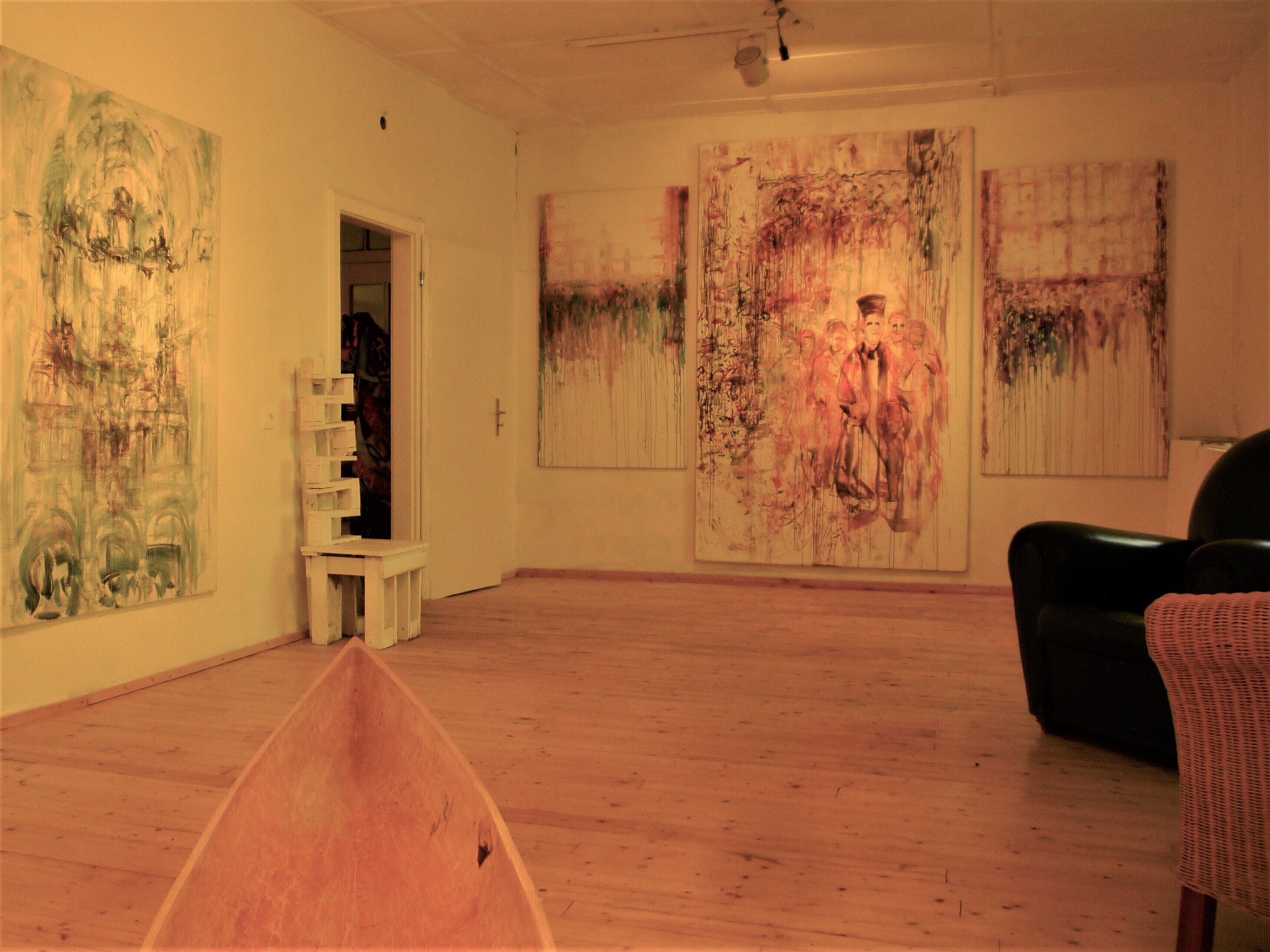 Une galerie d’art avec de grandes peintures accrochées aux murs, un plancher en bois et une chaise noire.