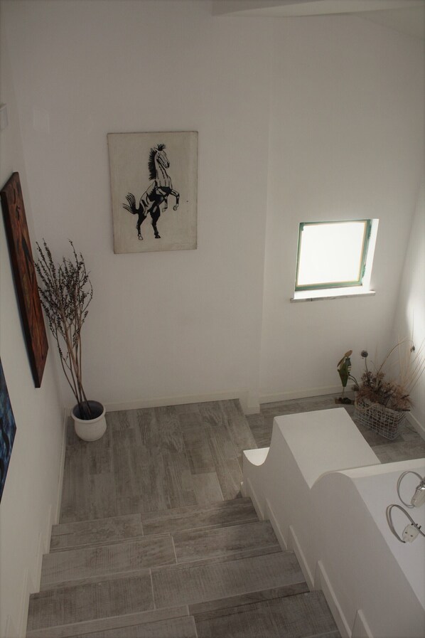 Un escalier recouvert de carreaux gris mène à une pièce blanche dotée d’une fenêtre et d’un tableau représentant un cheval.