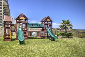 Children can play on the two slides and central platform of this playground, surrounded by a grassy area and a palm tree.