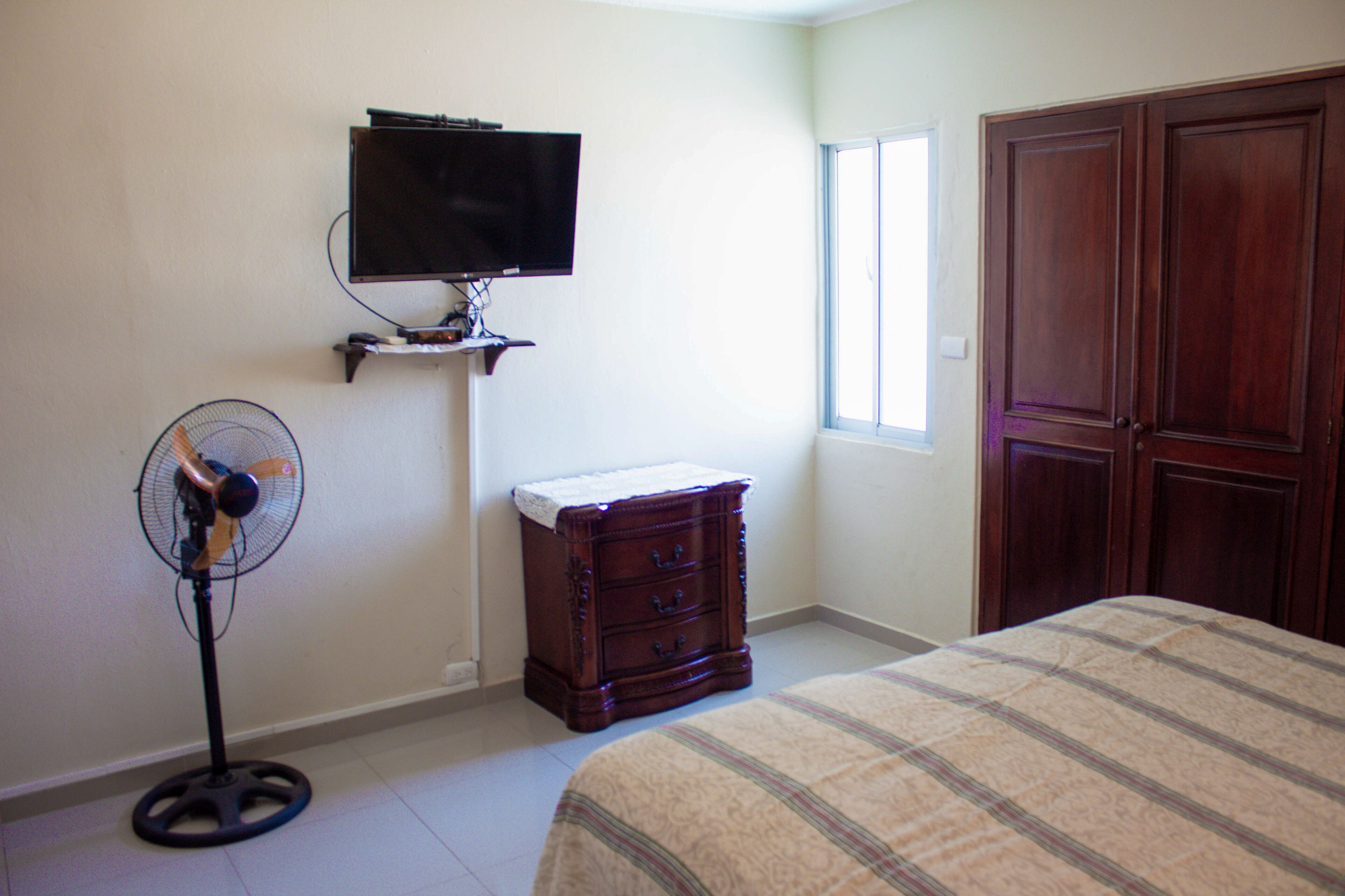 Une chambre à coucher avec un lit, une commode, un téléviseur posé sur un meuble, un ventilateur et une fenêtre.