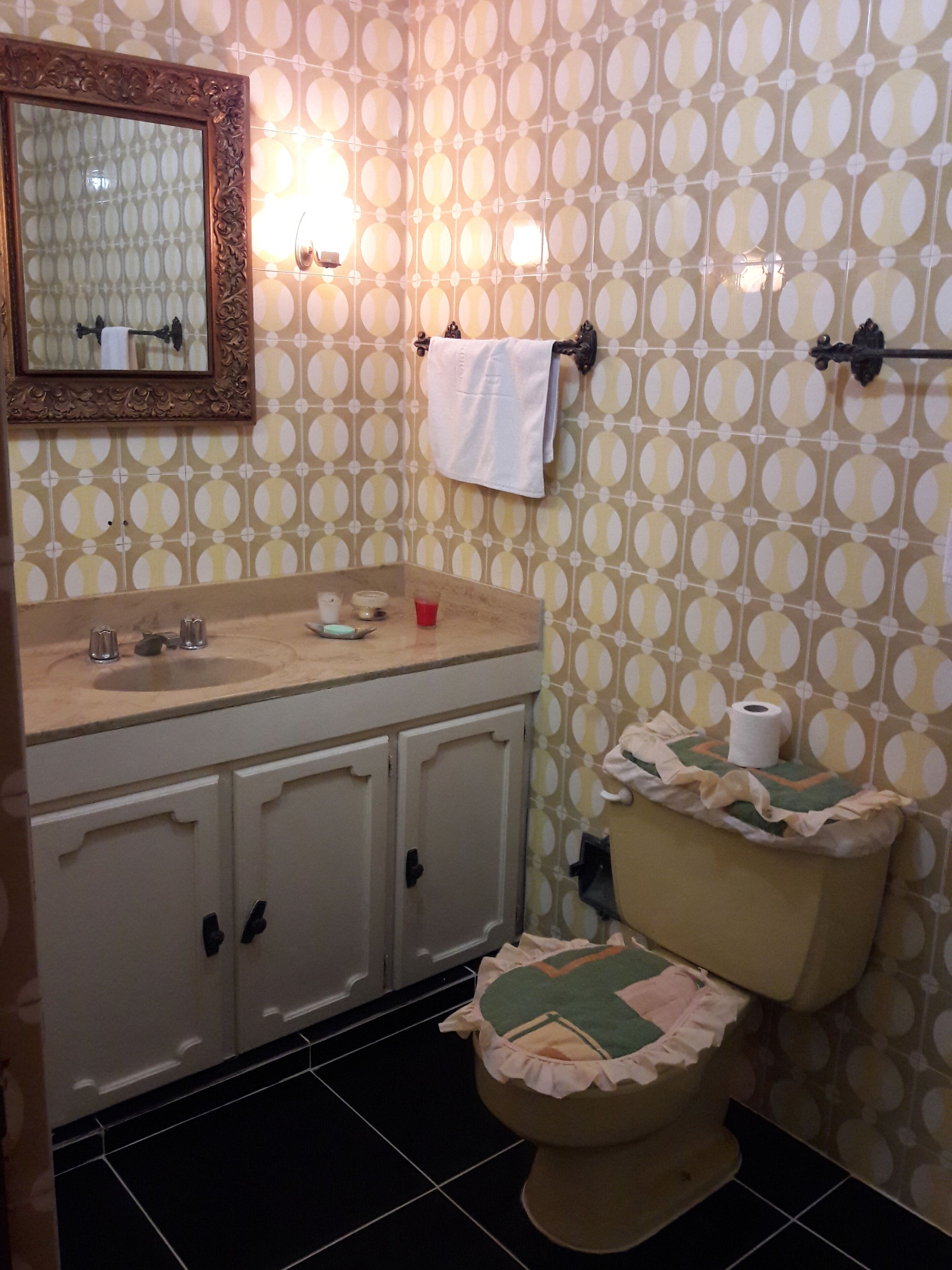 Un baño con pared de diseño, un lavamanos con artículos de aseo, un inodoro con cubierta decorativa y un espejo con marco de madera.
