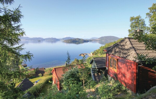 Et traditionelt træhus med tegltag, omgivet af frodig vegetation og udsigt over en stille sø med fjerne bjerge.