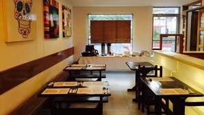 Interior de restaurante con mesas y bancos de madera, un amplio ventanal con vistas y coloridas obras de arte en las paredes.