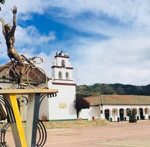 Une statue représentant une figure aux bras levés, placée sur un piédestal, un bâtiment blanc doté d’un clocher et une cour avec un toit en tuiles.