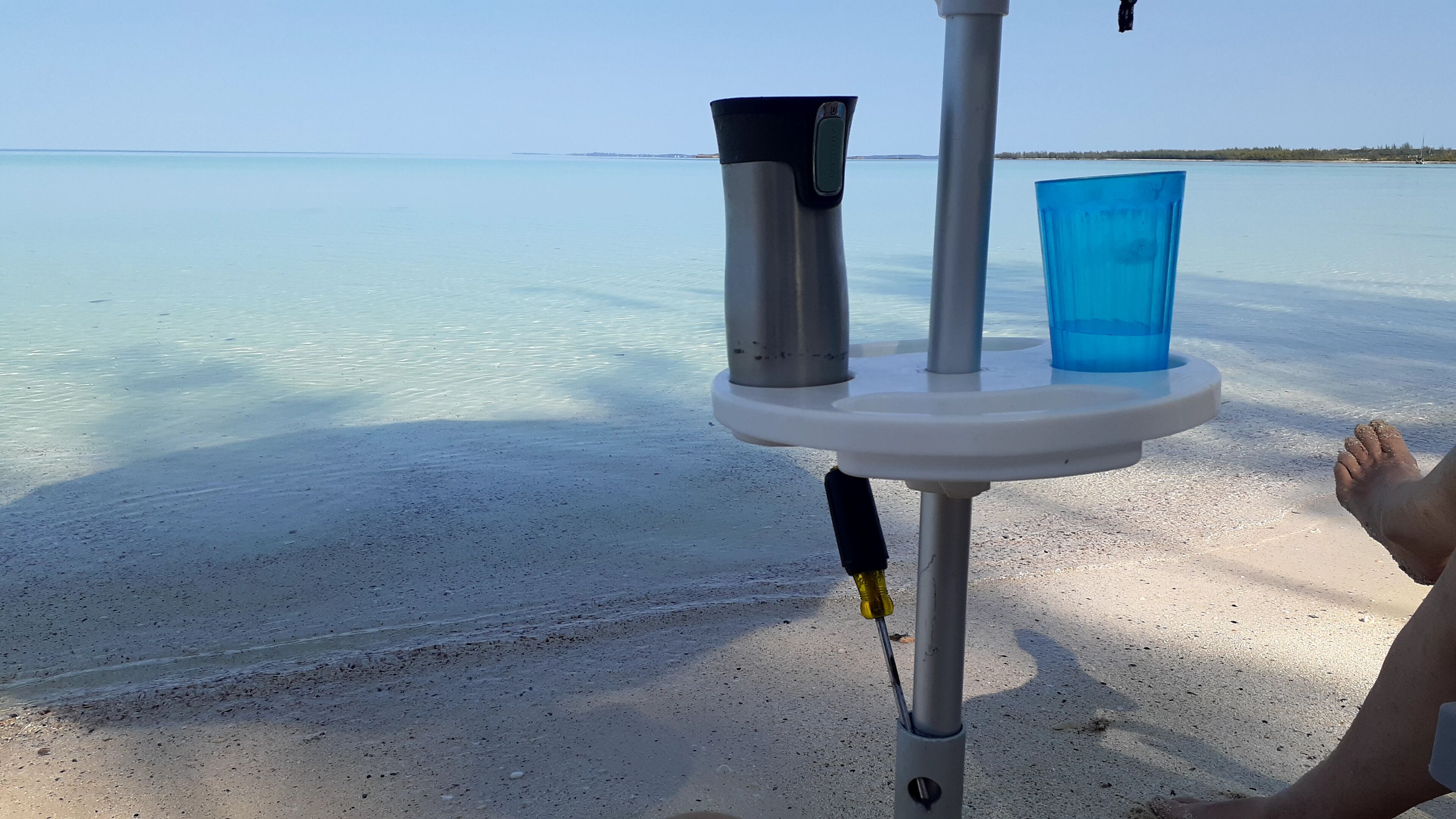 Een tafel aan het strand met een metalen beker en een blauwe plastic beker, een voet van een persoon, en een zandstrand met rustig water op de achtergrond.