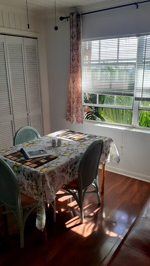 A dining area with a table covered by a floral tablecloth, two chairs, a window with blinds, and a view of greenery outside.
