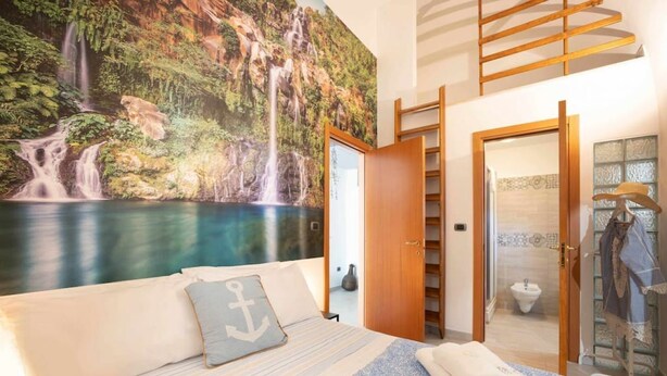 Een slaapkamer met een bed, een muurschildering van een waterval, een houten ladder en een zichtbare badkamer door een geopende deur.