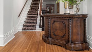 A grand staircase with a wooden finish, flanked by white walls and a dark wooden console table with a vase of flowers. - The CAPE (Prince Edward)