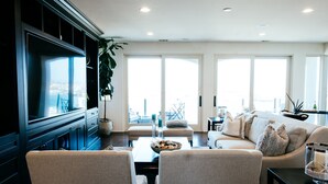 A modern living room with a large TV, comfortable seating, a coffee table, and large windows offering a view of the city. - Gorgeous Spanish-style Home with Stunning Ocean Views. New Stil™ jacuzzi! (Newport Beach)