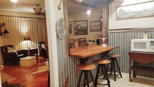 A cozy living room with a wooden dining table, bar stools, and a black leather chair. A white microwave is on a wooden stand in the kitchen area. - Unique Western Flair, Close to EXTRACO,  SILOS, Shopping, EATERIES, 3min LAKE! (Waco)