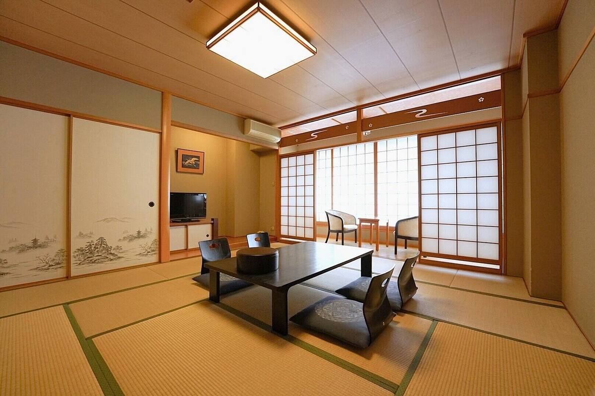 A traditional Japanese room with tatami flooring, a low table with cushions, sliding shoji doors, and a television on a shelf.