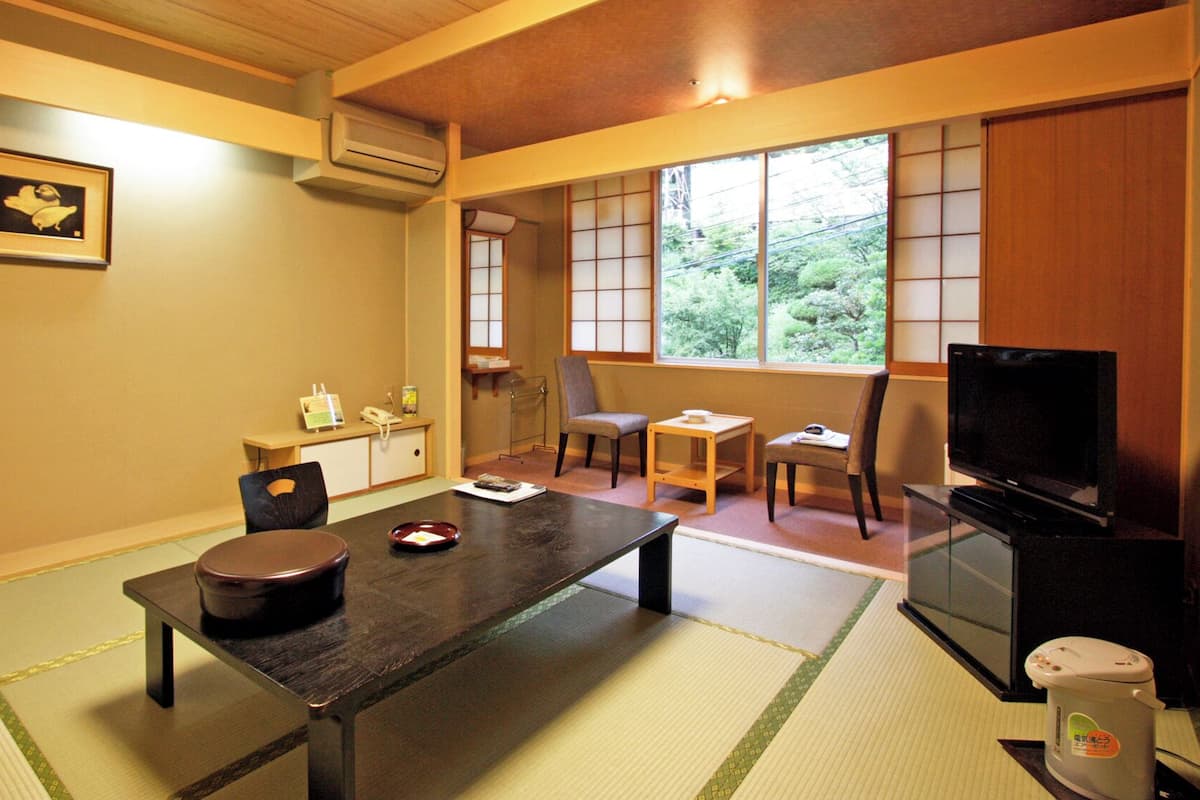 Ett traditionellt japanskt rum med tatamimattor på golvet, ett lågt bord med en mörk träskiva, en tv och en skjutdörr som leder ut till en utsikt över grönskan.