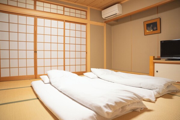Una habitación tradicional japonesa con suelo de tatami, futón, puertas correderas shoji y un televisor sobre un soporte de madera.