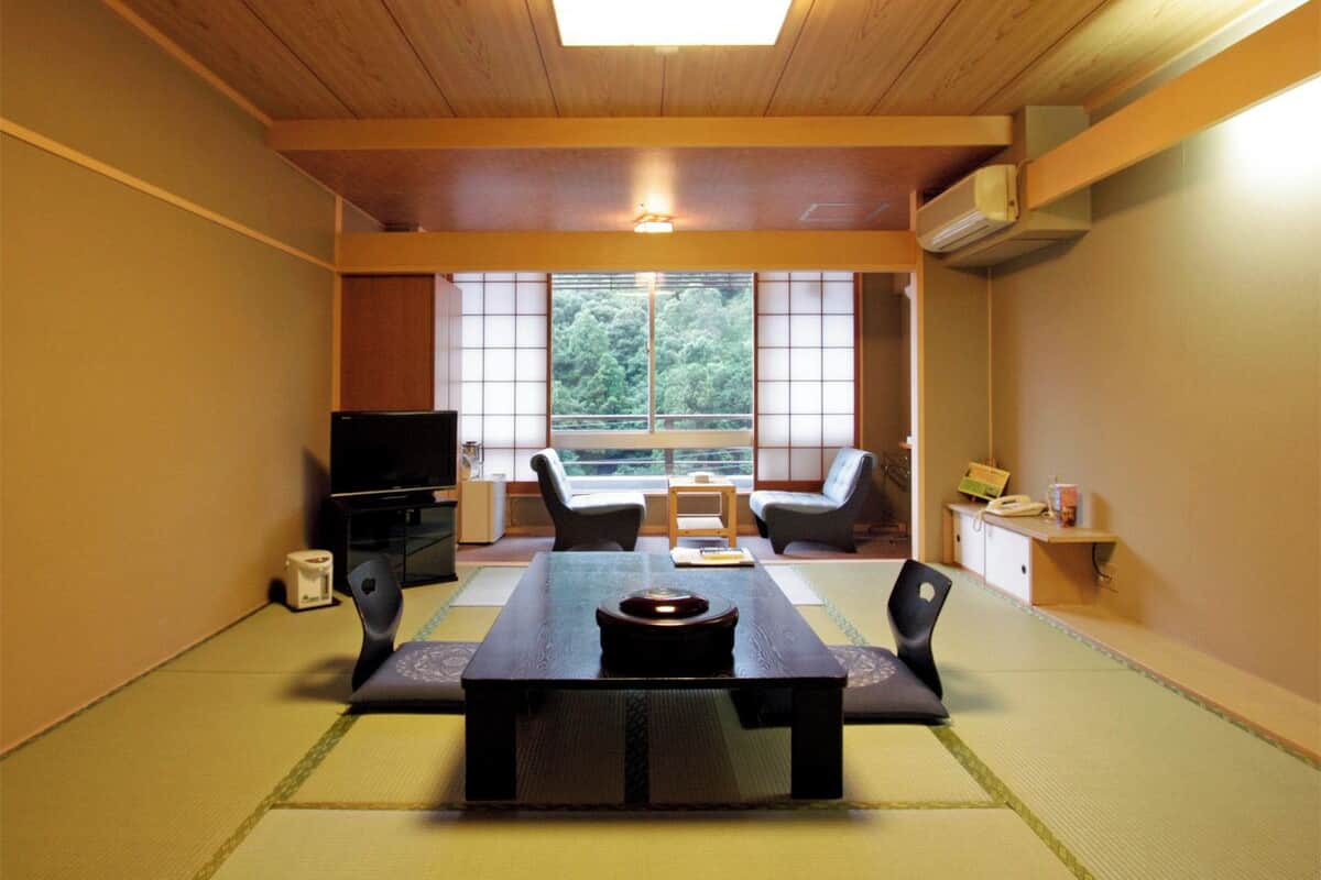 Ett traditionellt japanskt rum med tatamimattor på golvet, ett lågt bord, två stolar, en tv och en skjutdörr som leder ut till en balkong.