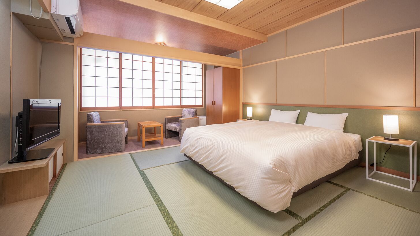 A hotel room with tatami mats, a bed, a television, a window with a view, and a traditional Japanese-style wall panel.