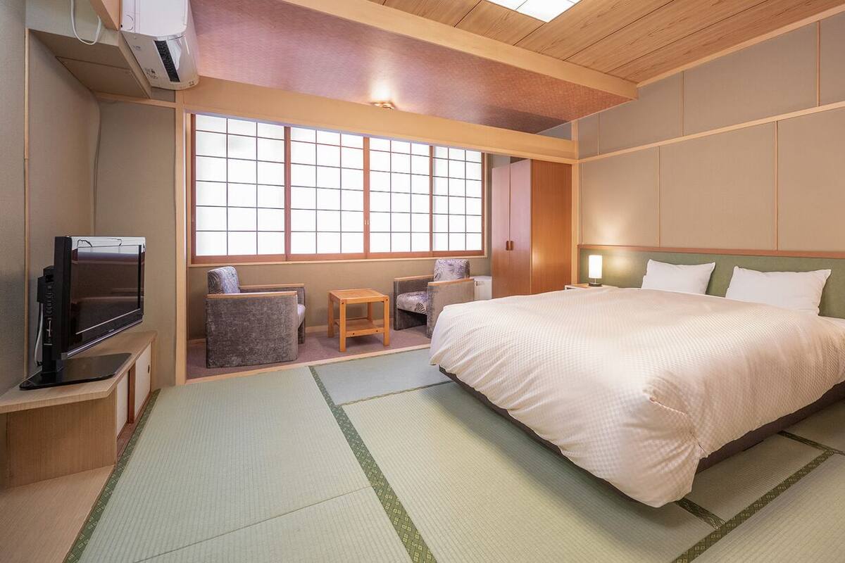 A hotel room with tatami mats, a bed, a television, a window with a view, and a traditional Japanese-style wall panel.