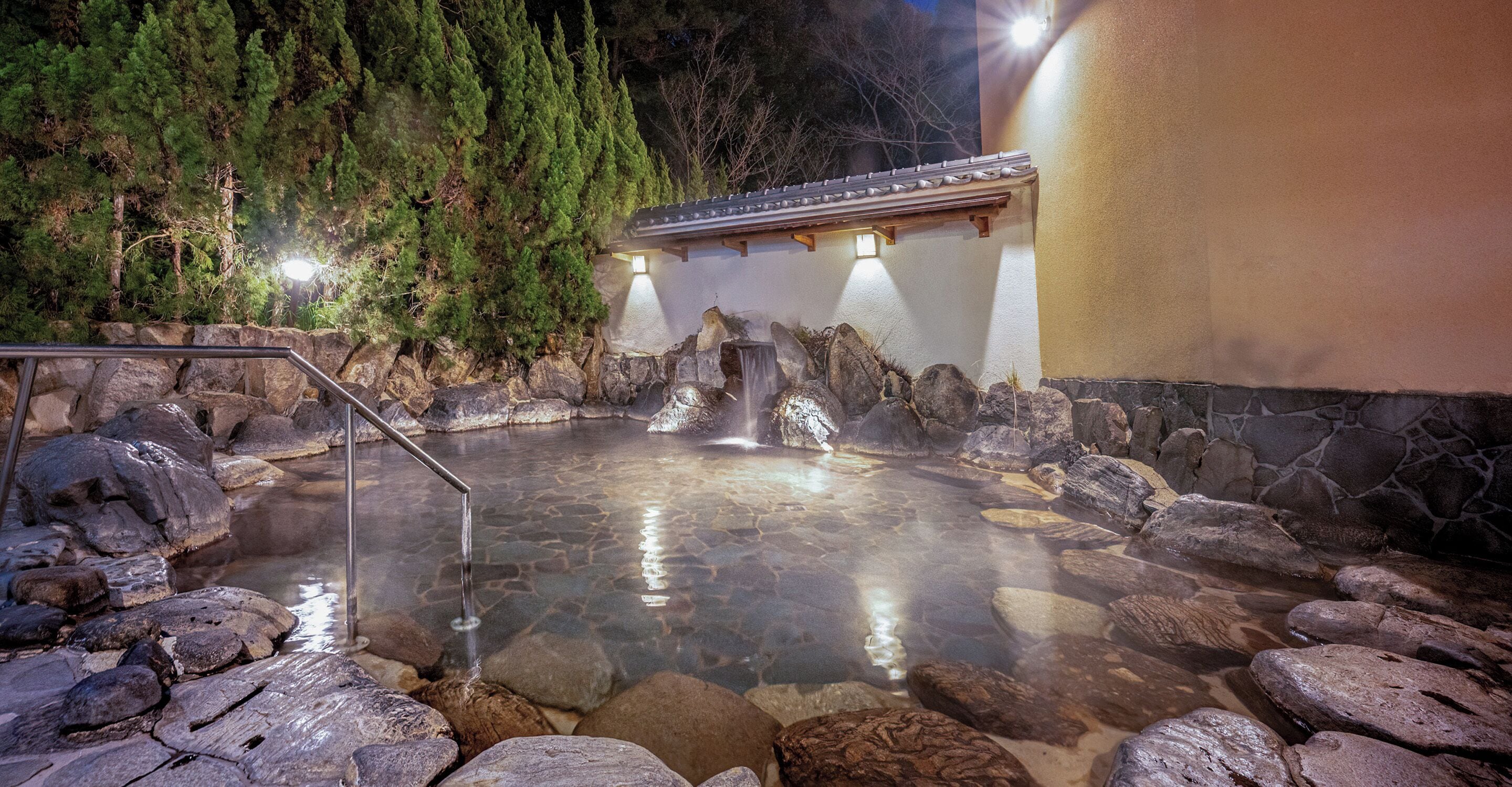 A hot spring area with a stone pool, a wooden building, and a waterfall.