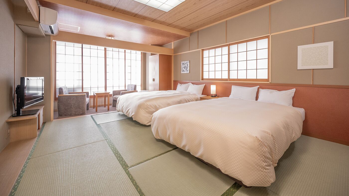 A hotel room with tatami mats, two beds, a desk with a TV, and large windows.