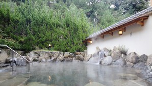 Un área de aguas termales con un camino de madera, rocas grandes y un edificio con tejado de baldosas.