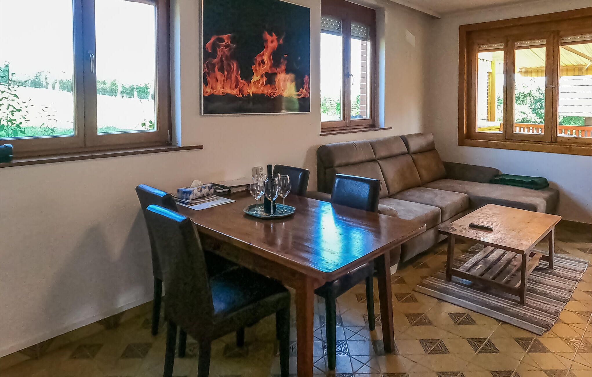 A dining area with a wooden table, chairs, and a sofa. There is a painting of flames on the wall and a window with a view of greenery outside.