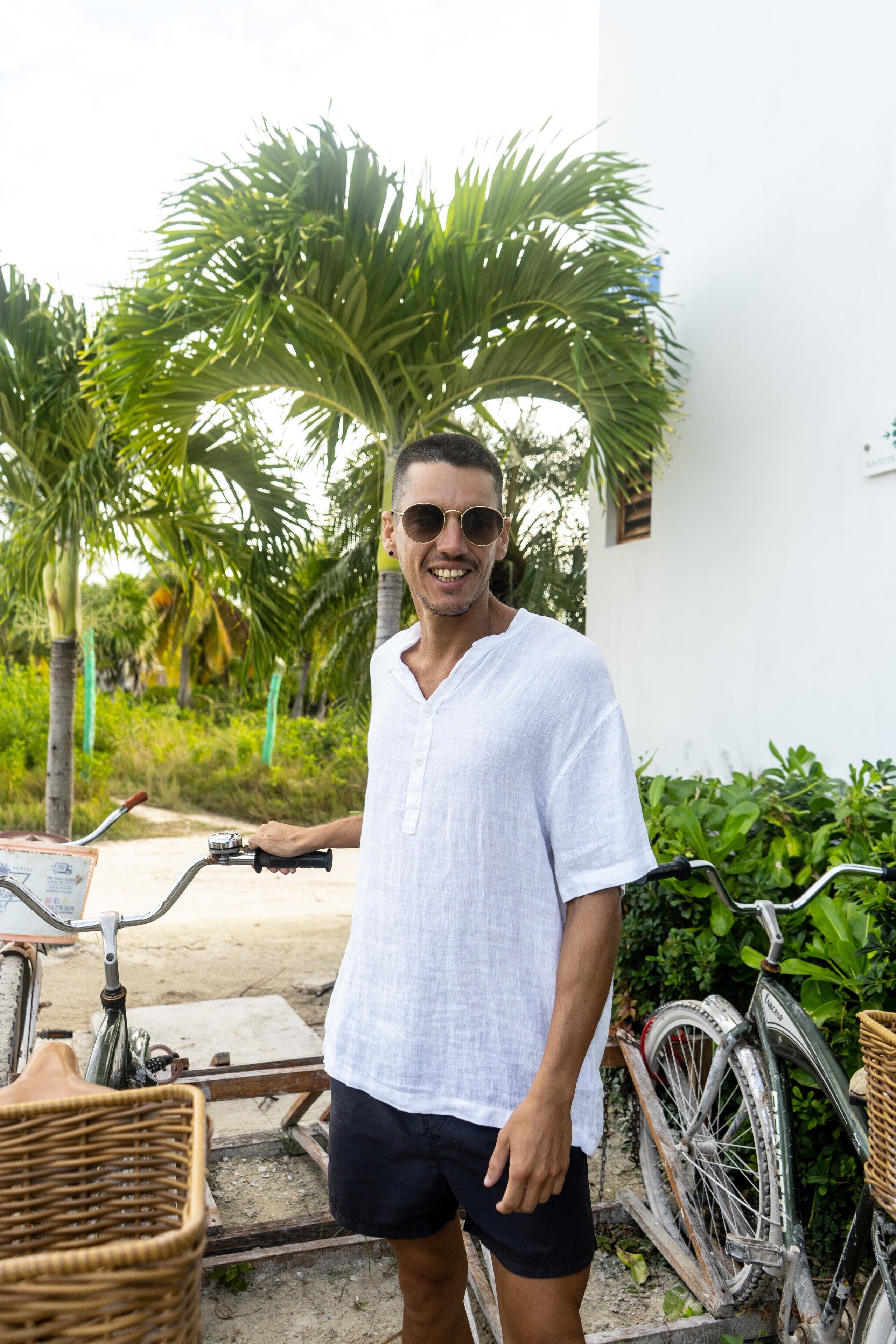 A man standing next to a bicycle with a basket, in front of a white building with greenery in the background.