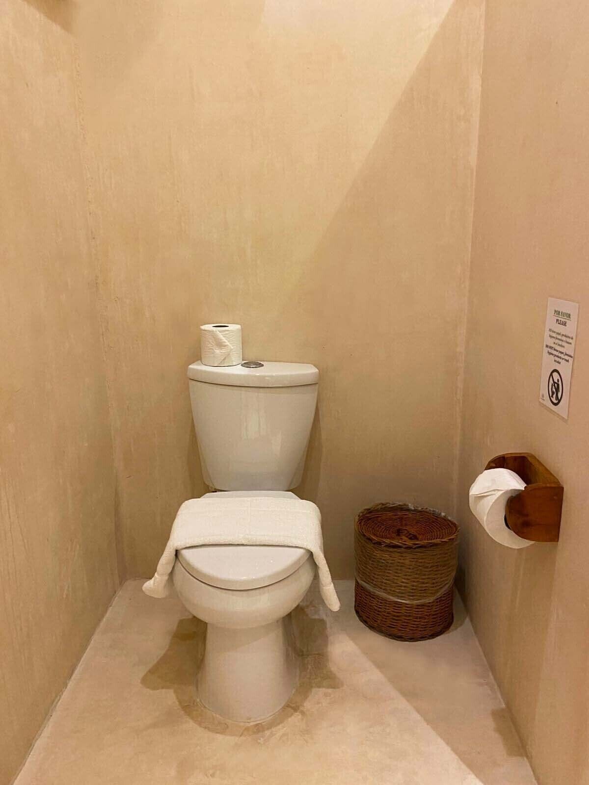 A small, compact bathroom with a toilet, a roll of toilet paper, a wicker basket, and a wall-mounted toilet paper holder.