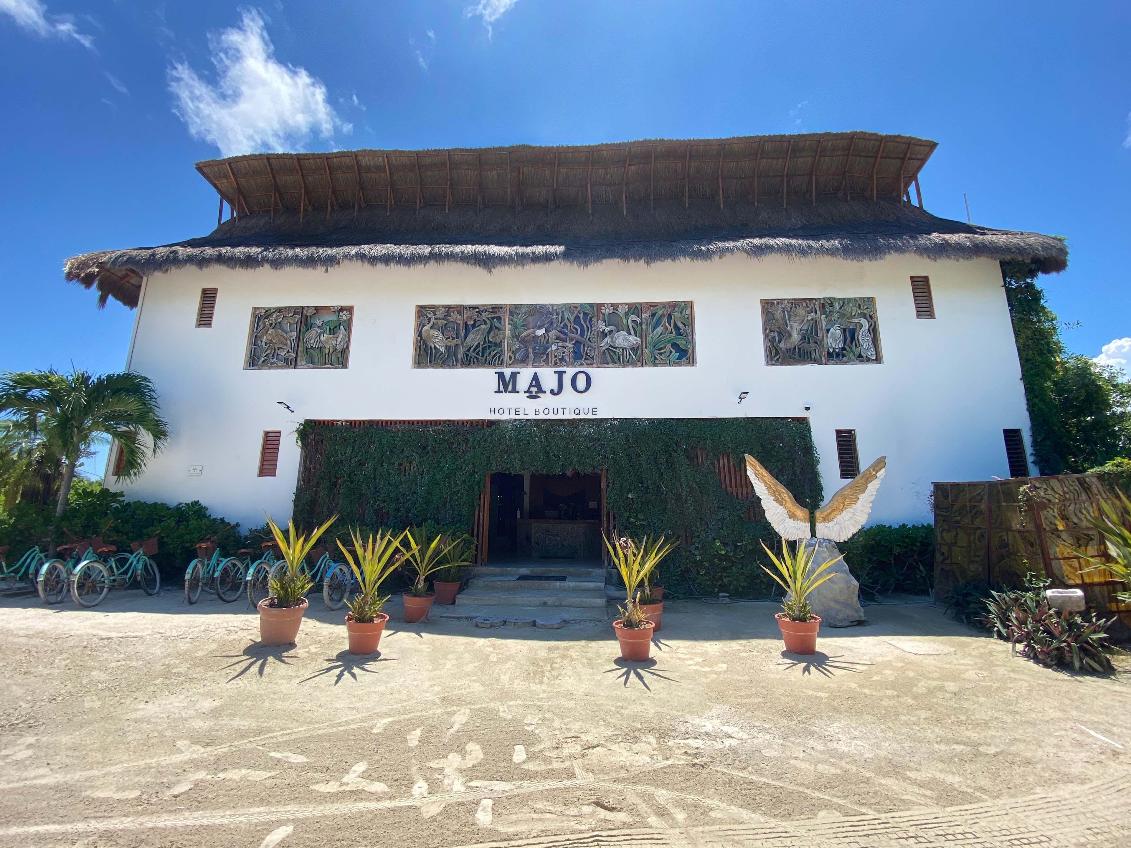 Un hotel llamado MAJO con techo de paja, un letrero y plantas en macetas en la entrada.