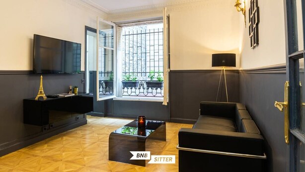 A modern living room with a black sofa, a coffee table, and a TV on a black stand. There is a window with a view of a balcony with plants. - MAGNIFICENT APARTMENT NEAR THE EIFFEL TOWER (Paris)