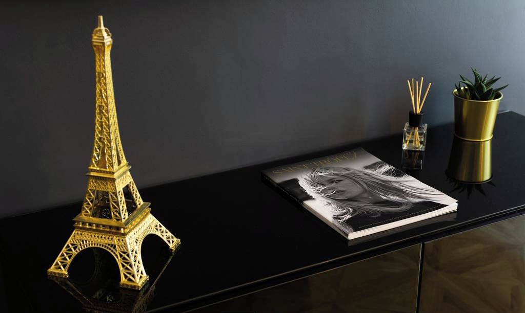 Un escritorio negro con un modelo dorado de la Torre Eiffel, un libro, una planta pequeña en maceta y un frasco de perfume.
