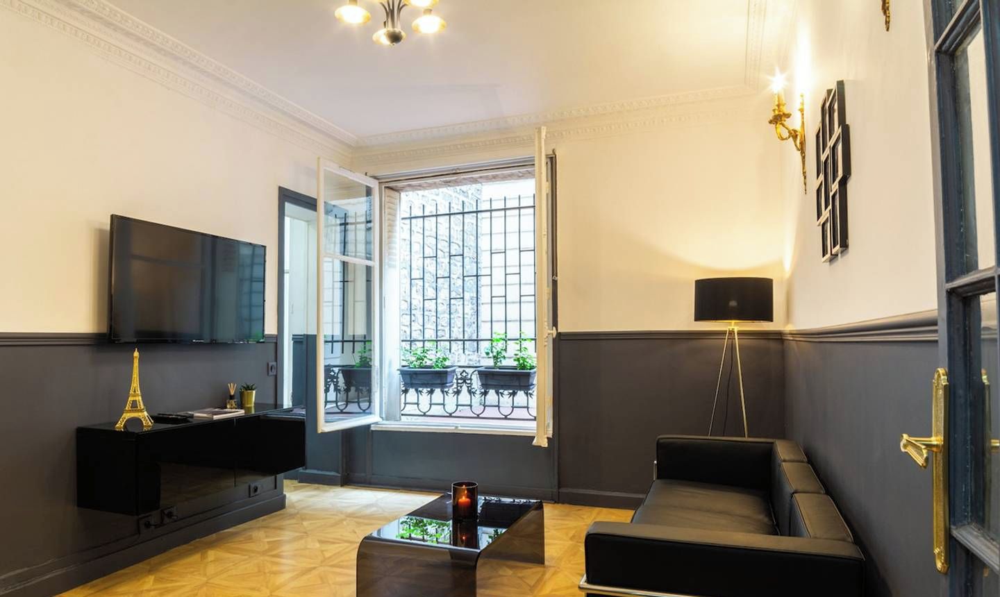 Un salon moderne avec un canapé noir, une table basse et un téléviseur posé sur un meuble noir. Une fenêtre donne sur un balcon agrémenté de plantes.