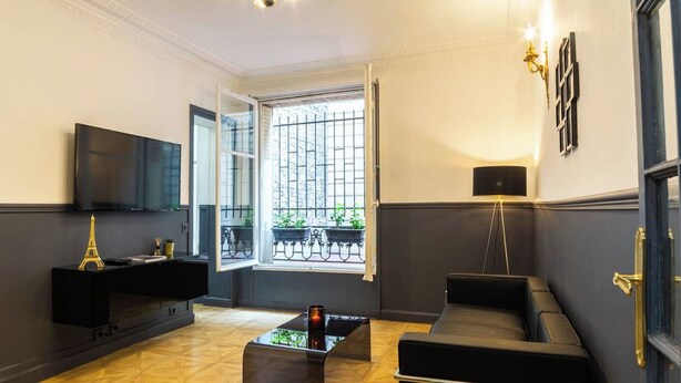 A modern living room with a black sofa, a coffee table, and a TV on a black stand. There is a window with a view of a balcony with plants.