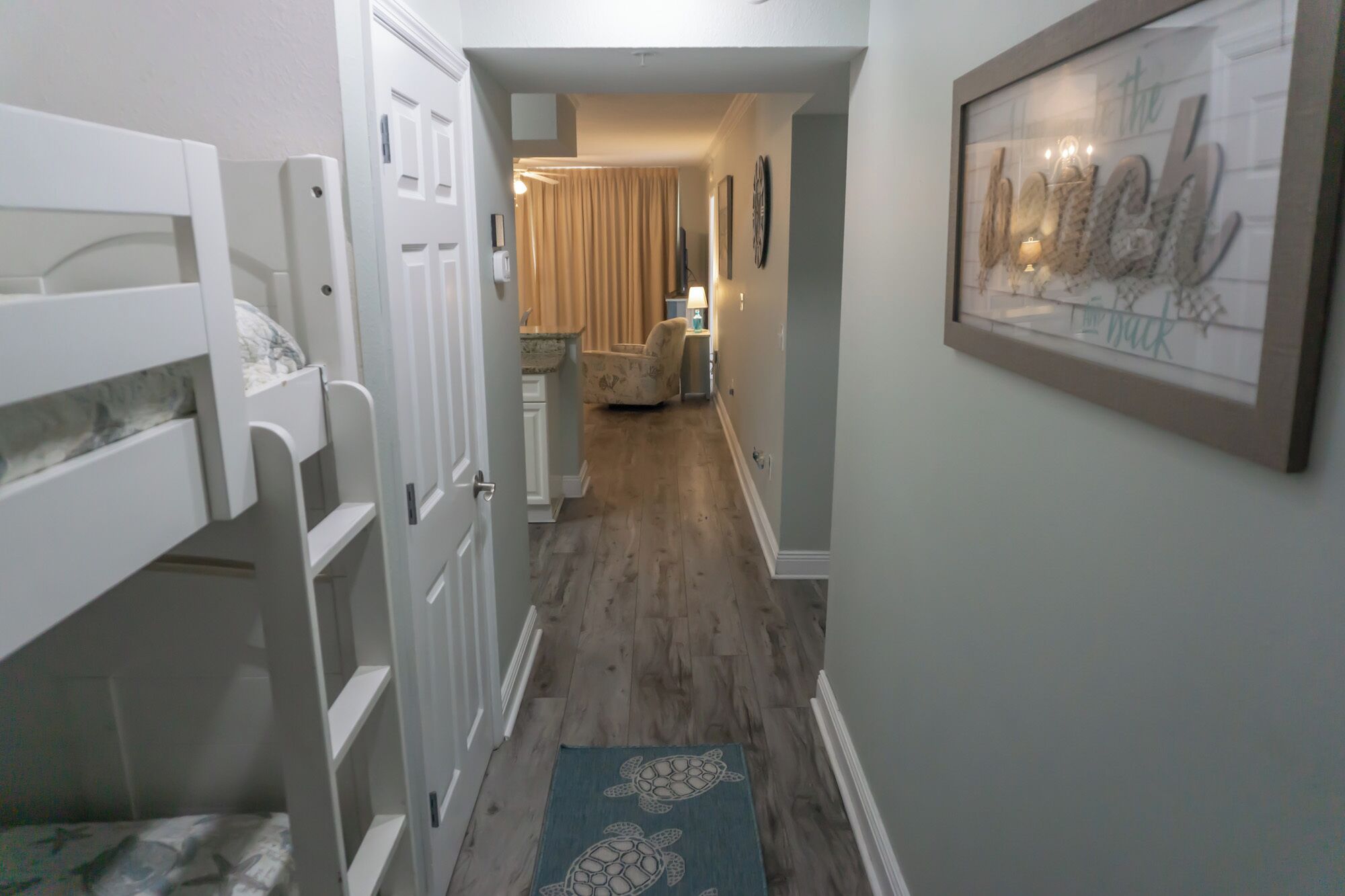 A narrow hallway with a wooden floor, a blue rug with turtle designs, a white door, and a framed picture on the wall.