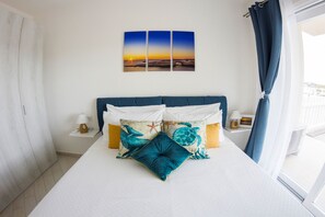 Hotellihuone, jossa on sänky, kaksi yöpöytää, ikkuna verhoilla ja seinä, jossa on kolme auringonlaskukuvaa.