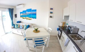 Una cocina luminosa y moderna con armarios blancos, un lavamanos de acero inoxidable y un microondas. Hay un comedor con mesa blanca y sillas azules, y un sofá con cojines de colores.