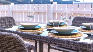 Une table à manger dressée avec des assiettes, des bols et des verres, entourée de chaises en osier, avec une clôture blanche et des montagnes en arrière-plan.