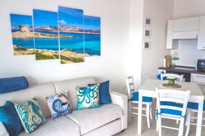Una sala moderna con un sofá color beige, sillas azules y blancas, y una mesa de comedor. Se observa un cuadro grande que representa un paisaje costero.
