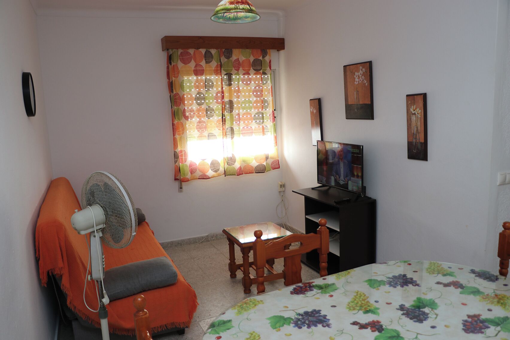 A bedroom with a bed, a desk, a chair, a standing fan, a television, and a window with curtains.