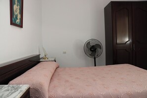 Ein Schlafzimmer mit einem Bett, einem Schreibtisch, einem Stuhl, einem Standventilator und einem Kleiderschrank.