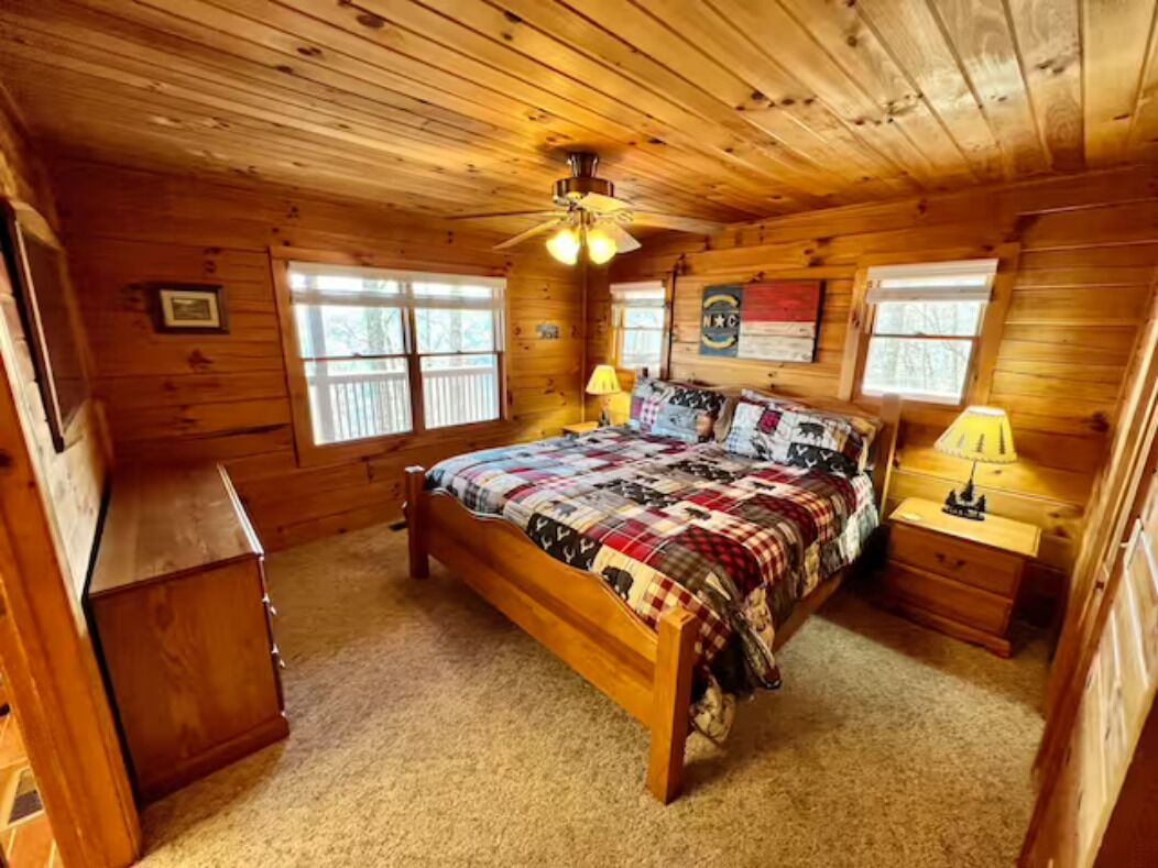 Une chambre dans une cabane en bois, avec un lit, deux tables de chevet, un ventilateur de plafond et une fenêtre.