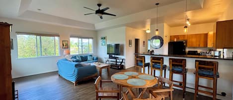 A spacious living area with a sofa, a dining table with chairs, a ceiling fan, and a kitchen area with wooden cabinets and a countertop.