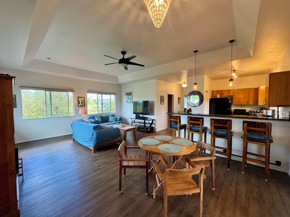 A spacious living area with a sofa, a dining table with chairs, a ceiling fan, and a kitchen area with wooden cabinets and a countertop.