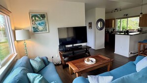A modern living room with a large TV, two blue sofas, a wooden coffee table, and a kitchen in the background. - Come & stay with us at the beautiful & peaceful Bird's Nest (Keaau)