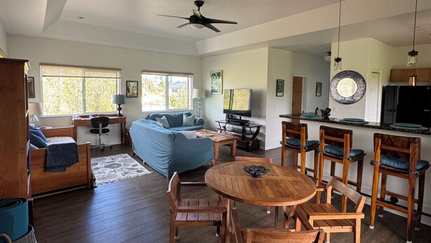 A spacious living room with a wooden dining table and chairs, a blue sofa, a ceiling fan, and a chandelier. - Come & stay with us at the beautiful & peaceful Bird's Nest (Keaau)